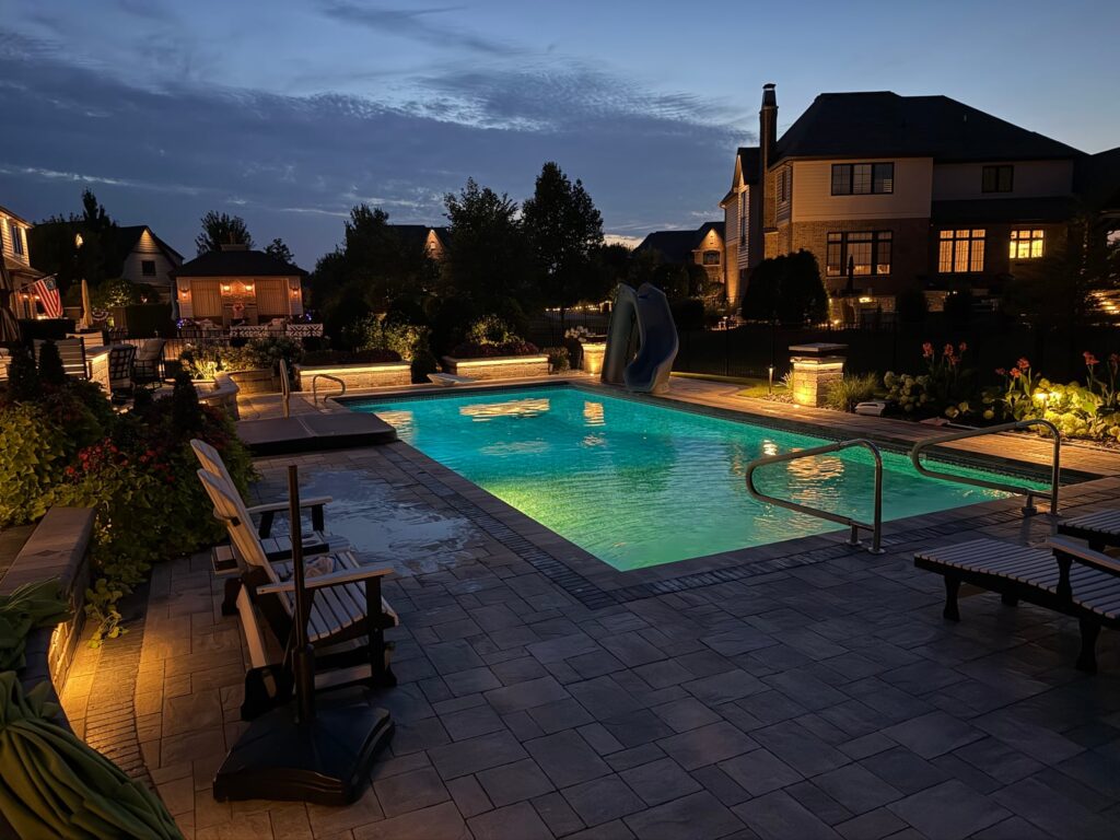 An elegant backyard pool area at dusk is warmly illuminated with linear silicone-injected white lighting along the planter boxes, outdoor kitchen grill and island, and the caps of multiple fire bowls, creating a soft, inviting glow around the water and seating areas