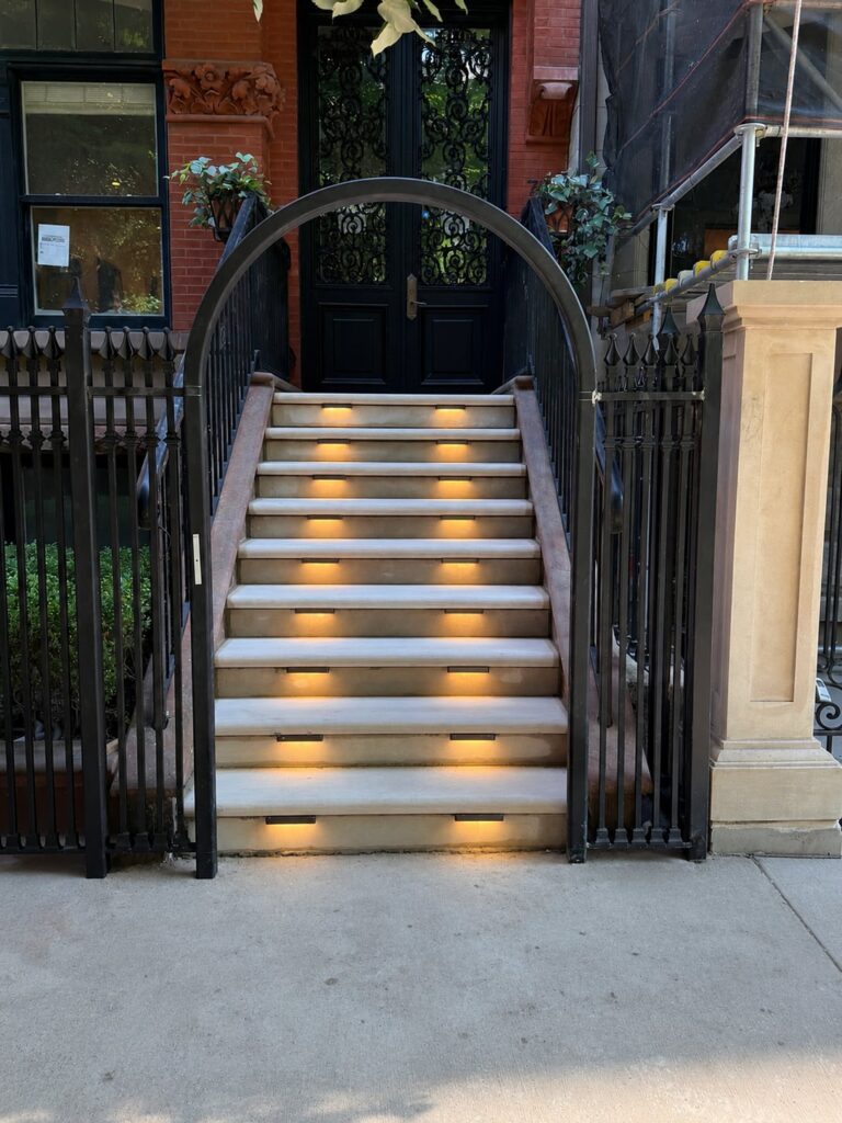 Warm brass LED step lights are neatly integrated into each riser of the front entry staircase, creating an inviting, evenly illuminated path up to the home’s ornate double doors
